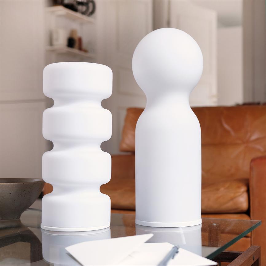 To hvide, skulpturelle vaser — én med bølgende segmenter, én med rund top — står på et glasbord foran en brun lædersofa i en lyst, minimalistisk opholdsrum.