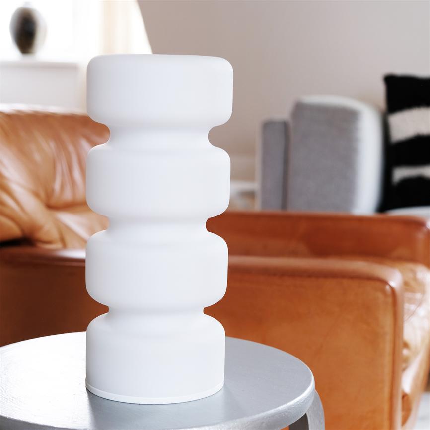 Hvid skulpturel bordlampe står på et rundt sølvfarvet bord foran en brun lædersofa og et gråt sofahjørne i et lyst, moderne opholdsrum.