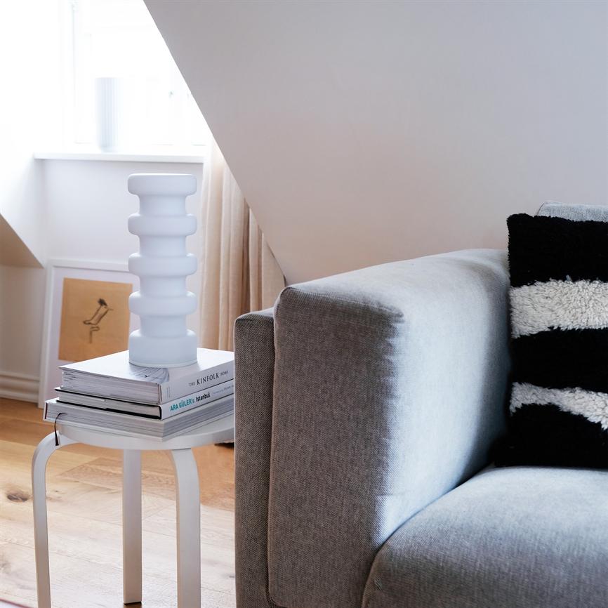 En hvid, rillede vase står på en stak bøger på et lille hvidt sidebord; ved siden af en grå sofa med sort-hvid pude under skrå loftsvæg og lys fra et vindue. 
 
 Transkription af synlig tekst: "THE KINFOLK HOME", "ARA GÜLER: Istanbul"