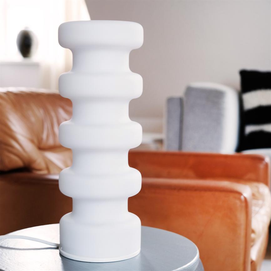 Hvid, skulpturel bordlampe står på et rundt sidebord; den bølgede cylinderform kontrasterer brune læderstole og grå polstrede møbler i et lyst, moderne stueindretning.