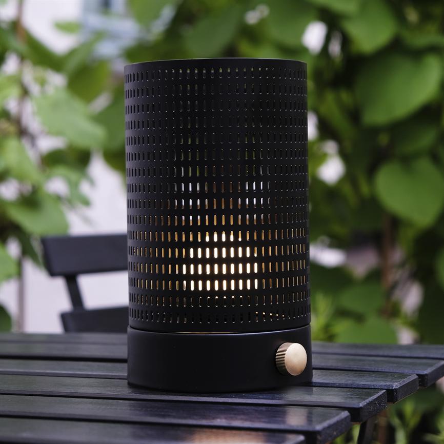 Sort perforeret cylindrisk bordlampe lyser varmt gennem små huller, stående på et mørkt træbord på en balkon med grønt løv i baggrunden.