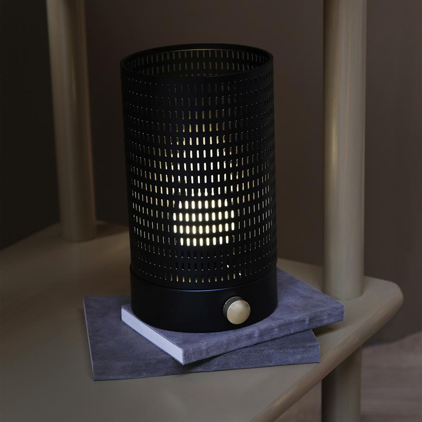 Cylindrisk sort metal lampe med guldfarvet drejeknap udsender blødt lys gennem perforerede huller; den står på to grå stofindbundne bøger på en beige træhylde med lodrette stolper.