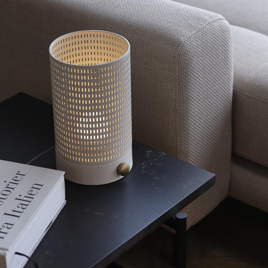 En cylindrisk, perforeret bordlampe udsender varmt, gennemlyst skær, stående på et mørkt sidebord ved en beige sofa ved siden af en bog mærket "Historier fra Italien".