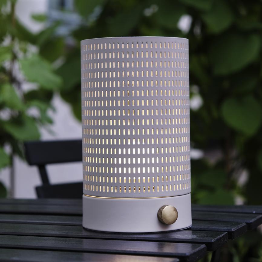 En cylinderformet lampe i perforeret metal lyser varmt gennem rektangulære huller; messingknap ved bunden, placeret på et sort lamelbord udendørs med sløret grøn bladbaggrund.