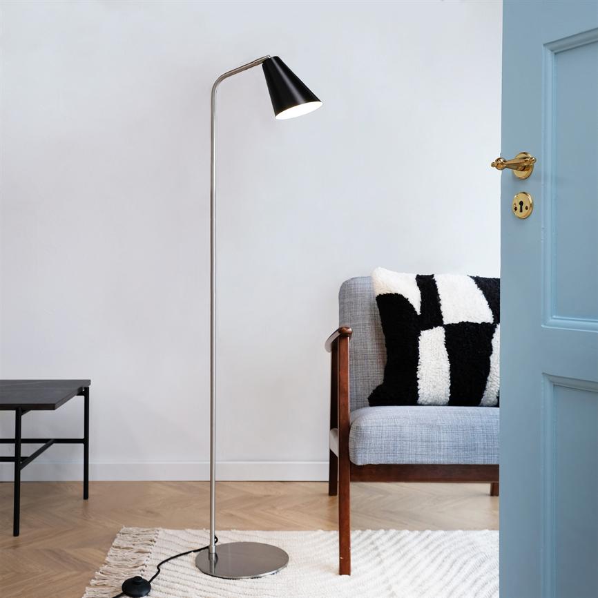Gulvlampe står og kaster varmt lys nedad ved en grå sofa med en sort-hvid pude; åben lyseblå dør, lille sort sidebord, lyst tæppe og trægulv i et roligt hjørne.
