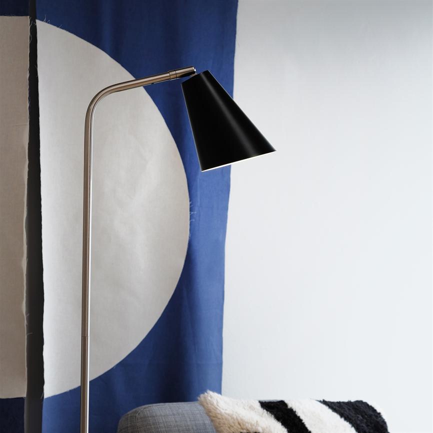 Gulvlampe med sort kegleskærm bøjer sig over en grå sofa; den lyser ned foran et blå-hvidt gardin med stort cirkelmotiv, ved en sort-hvid pude.