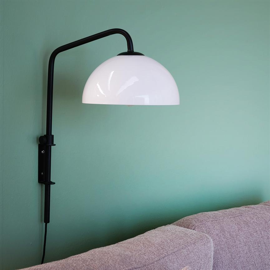 Væglampe med sort arm og hvid rund skærm hænger over ryglænet på en lyserød sofa mod en mintgrøn væg, kablet løber ned langs væggen.