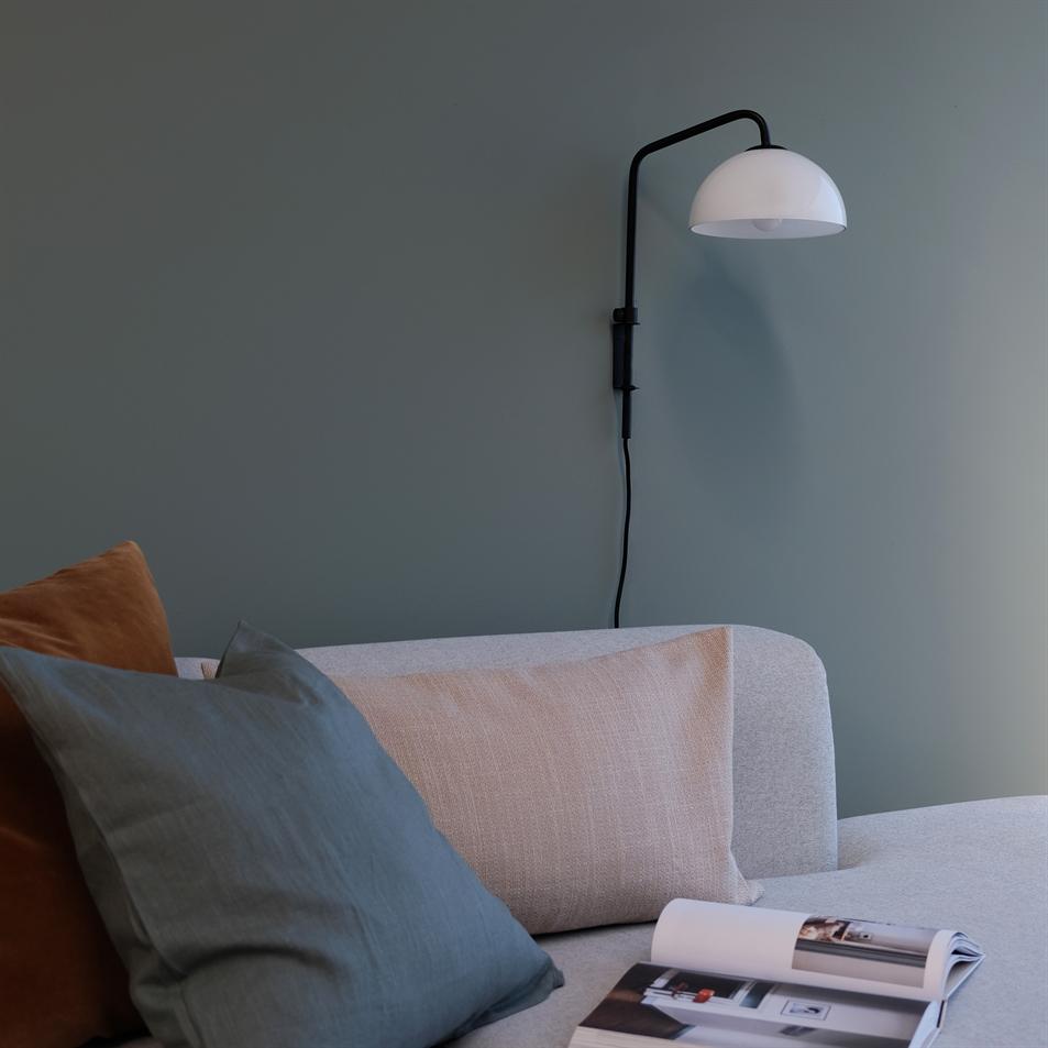 Sofaen i lysegråt med to farvede puder, et åbent magasin ligger på sædet; en sort væglampe bøjer ned mod sofaen, alt foran en afdæmpet støvet grøn væg.