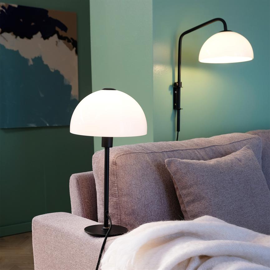 En hvid kuppelbordlampe står og lyser på armlænet af en beige sofa; en tilsvarende væglampe kaster lys mod en mintgrøn væg med et abstrakt maleri i et stuehjørne.