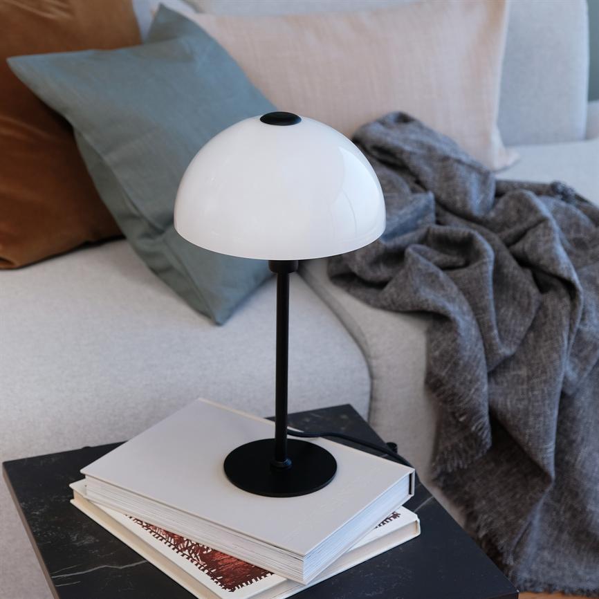 Bordlampe med hvid kuppel på sort stang hviler ovenpå en stak bøger på et lille sort bord; ved siden af en lys sofa med farvede puder og et gråt tæppe.