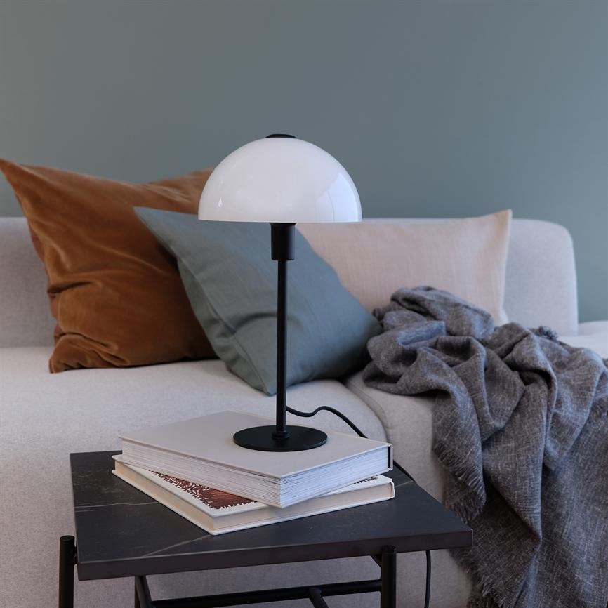 En bordlampe står på en stak bøger på et lille bord; i baggrunden en lys sofa med brune og grønne puder og et gråt sammenkrøllet tæppe foran en blågrå væg.