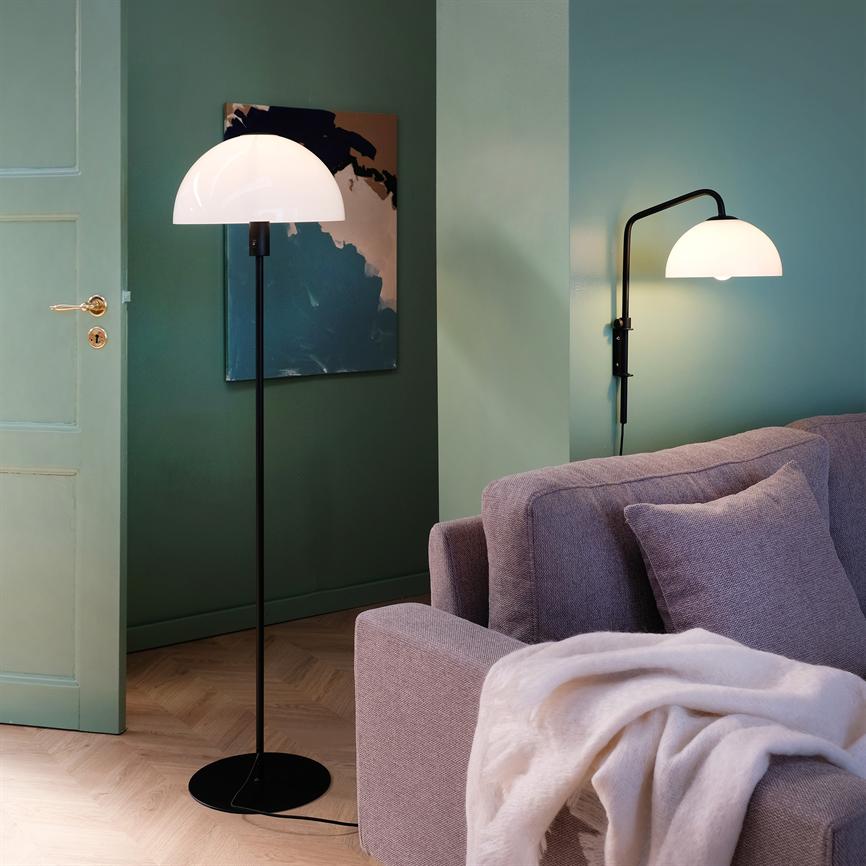 En sort gulvlampe med halvkugleformet hvid skærm lyser blødt; en matchende væglampe over en lysegrå sofa med tæppe, placeret mod grønne vægge og et abstrakt maleri.