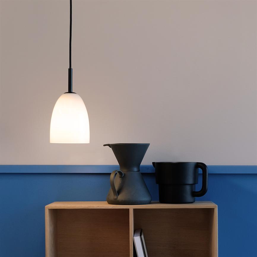 En hvid, kuppelformet hængelampe lyser blødt over en lille træhylde, hvor en sort, tragtformet kaffekande og et sort krus står mod en blå og beige væg.