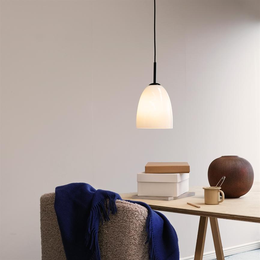 En hængende hvid lampe lyser over et lyst træbord; på bordet ligger to æsker, en kop med ske og en brun keramikvase, foran en blød stol med blåt tæppe.