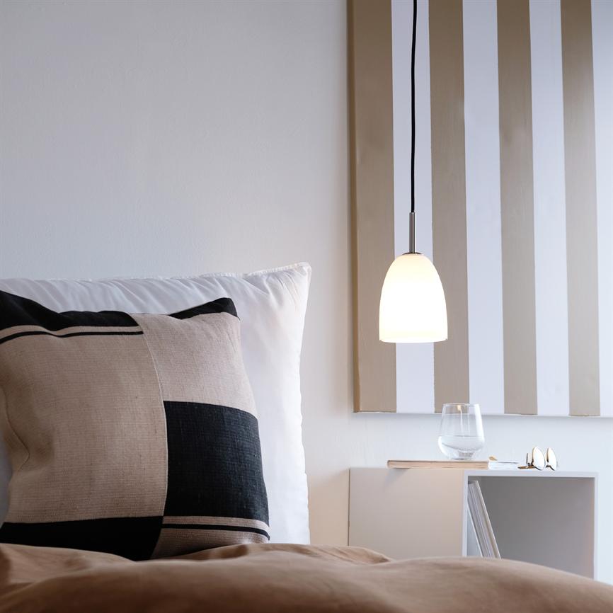 Lampe hænger og lyser over et natbord; ved siden sidder en stor ternet pude på en seng, og natbordet rummer et vandglas, briller og en stribet vægdekoration.