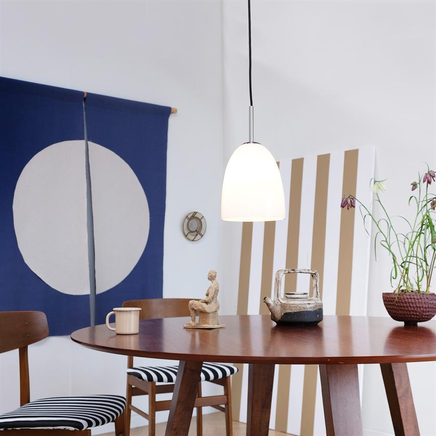 En hvid hængelampe hænger over et rundt træbord, hvor en lille skulptur, tekande, kop og plante står; træstole med sort-hvide stribede hynder i et lyst, minimalistisk rum.