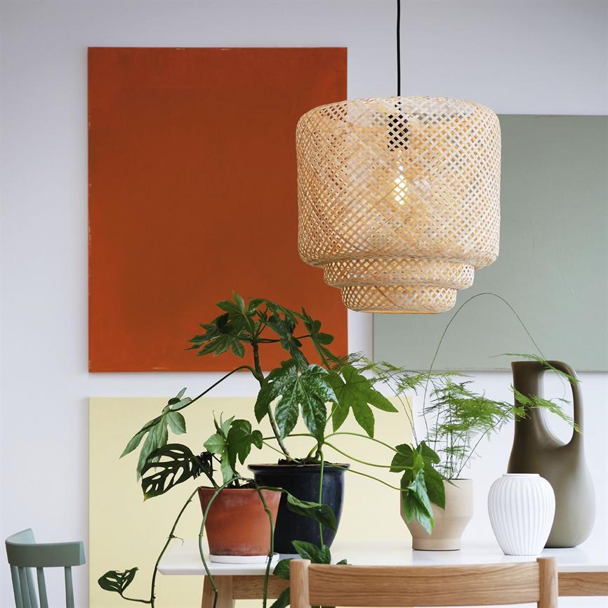 En flettet lampe hænger og lyser over et bord; flere stueplanter i keramikpotter står på bordet foran farvede maleriplader på væggen i et lyst spiseområde.