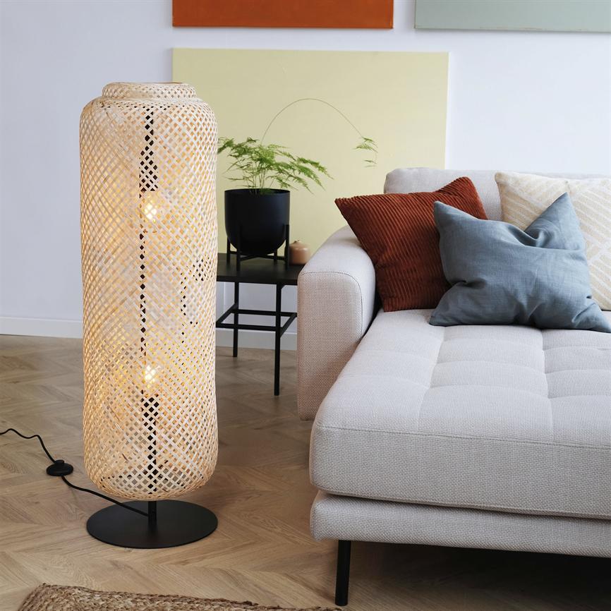En høj, flettet gulvlampe lyser blødt; den står ved en lysgrå sofa med rustfarvede og blå puder, foran et sort sidebord med plante på lys parket og vægkunst.