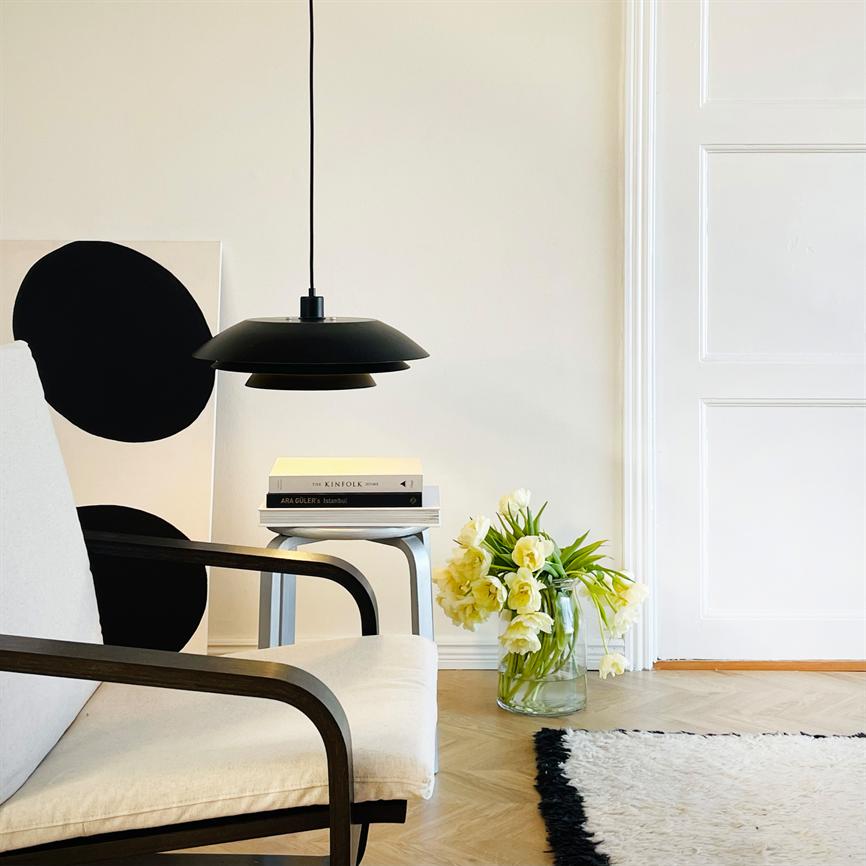 Sort loftlampe hænger over et lille sidebord med bøger i en lys, minimalistisk stue med en beige armstol, trægulv, hvid dør og en glasvase med gule blomster.
 Tekst: THE KINFOLK TABLE; ARA GULER - Istanbul