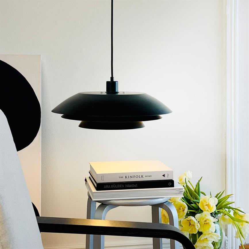 Sort pendel-lampe hænger over en stabel bøger, kaster blødt lys; bøgerne står på en lille skammel ved en stol og en buket gule tulipaner i et lyst rum.
 
 Tekst i billedet: THE KINFOLK HOME; ARA GÜLER'S Istanbul