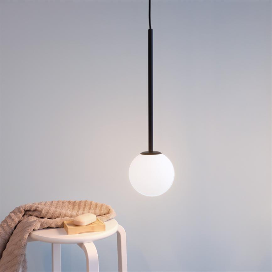 En rund, hvid pendel lyser blødt; den hænger over et lille hvidt bord, hvor et foldet beige tæppe og en sæbe på en træbakke ligger i et lyst, minimalistisk rum.