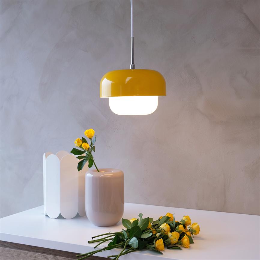 Gul loftlampe hænger tændt over en hvid bordplade med en beige vase og gule roser, foran en grå, struktureret væg.