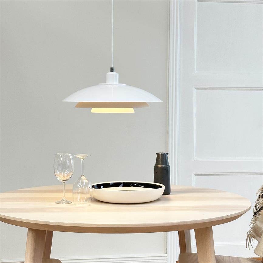 En hvid hængelampe hænger over et rundt lyst træbord, hvor en hvid skål, to vinglas (et vendt på hovedet) og en mørk kande står i et lyst, minimalistisk rum.