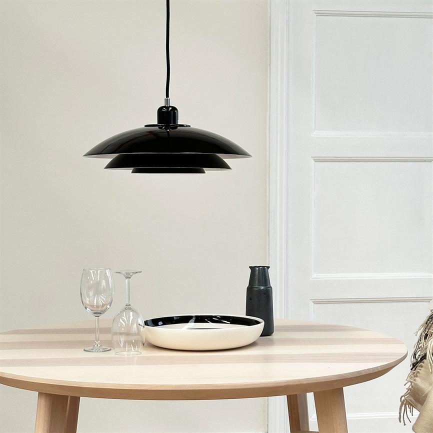 Sort pendellampe hænger over et rundt lyst bord; en sort-hvid skål, to vinglas (et opret, et på hovedet) og en sort kande står foran en hvid væg med dør.