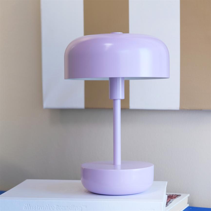 En lilla, kuppelformet bordlampe står på en bunke bøger foran en beige-hvid stribet vægdekoration i et neutralt rum. Tekst på bogens kant: "illustrativeBranding™".