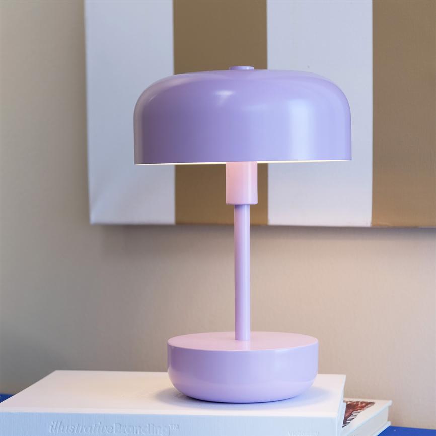 En lilla bordlampe står tændt og kaster blødt lys nedad, placeret på en hvid bog på et bord foran en beige væg med brede hvide og brune striber. 
 Tekst: "illustrativeBranding™"