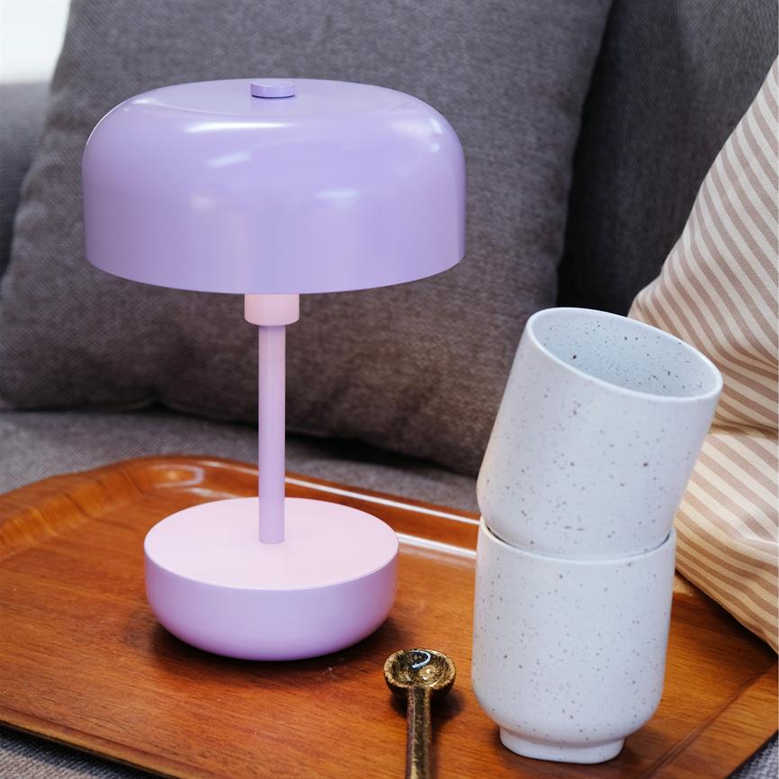 Lilla bordlampe står på en træbakke, to hvide stænkede krus er stablet og lænet mod hinanden, en lille messingske ligger foran, alt placeret på en grå sofa med puder.