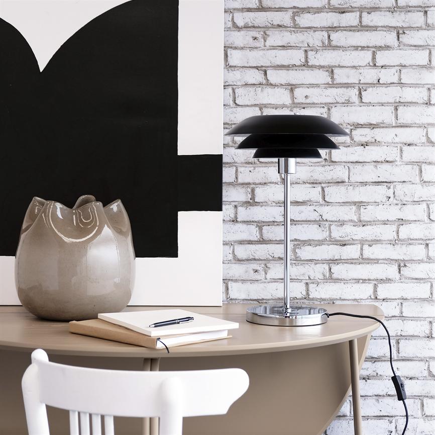 Sort, flerlaget bordlampe står midt på et rundt beige skrivebord; ved siden af ligger notesbøger og pen, en grå keramisk vase samt sort-hvidt abstrakt maleri foran hvid murstensvæg.