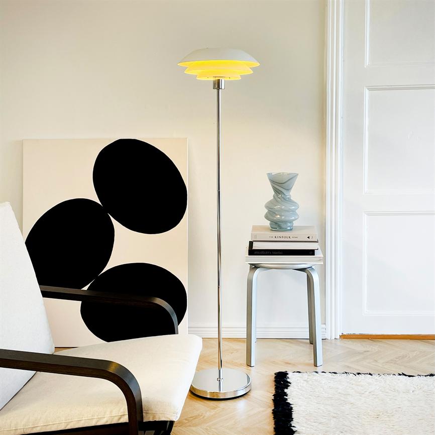 Gulvlampe står tændt ved en lys væg; ved siden af et lille bord med bogstak og keramisk vase; sofa og et abstrakt sort-hvidt lærred læner mod væggen. Ingen tydelig læsbar tekst.