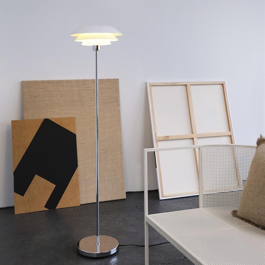 Gulvlampe står tændt og kaster blødt lys, ved siden af lænede lærreder og tomme rammer i et lyst, minimalistisk rum med en hvid metalstol og en pude.