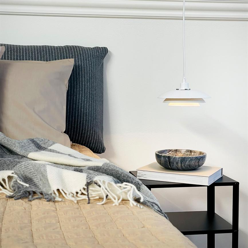 Hvid hængelampe lyser blødt over marmoreret skål og bog på et sort natbord; ved siden af står en seng med mørke og beige puder, ternet gråt tæppe og beige dyne.