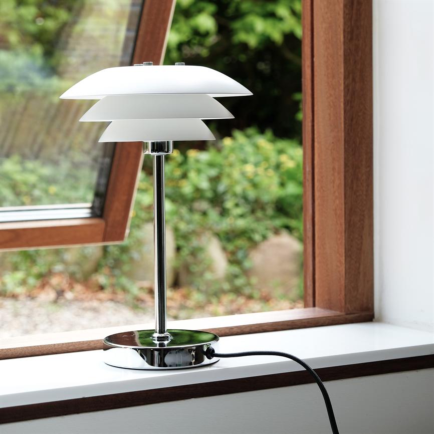 En hvid, lagdelt bordlampe står på en hvid vindueskarm, tilsluttet med sort ledning; et åbent trævindue indrammer en sløret grøn have udenfor.