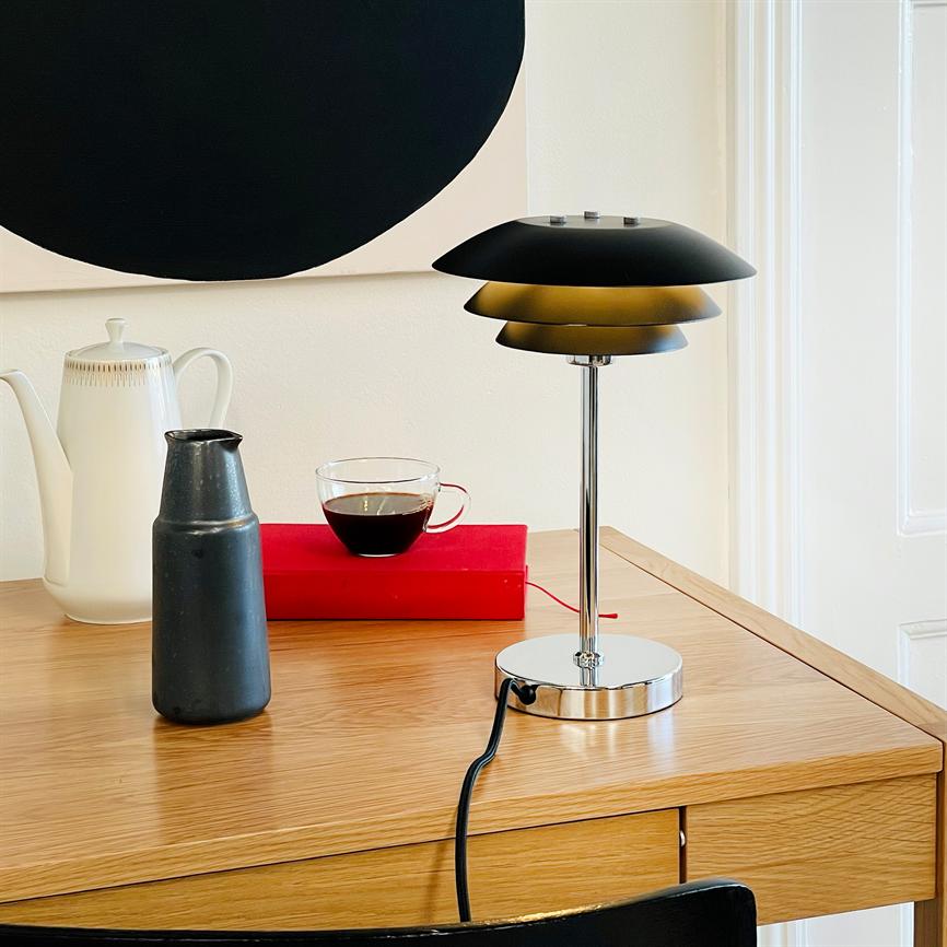 En sort-metallisk bordlampe står på et egetræsskrivebord ved en glas kop kaffe, en sort kande, en hvid kande og en rød bog foran en hvid væg med en sort cirkel.