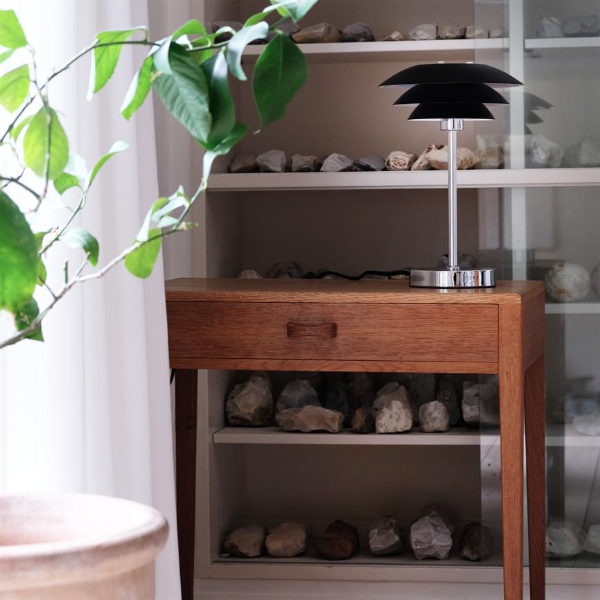 Et lille træbord med skuffe står mod en reol; en sort, moderne bordlampe står på bordets højre side, omgivet af hylder med sten og en grøn plante ved vinduet.