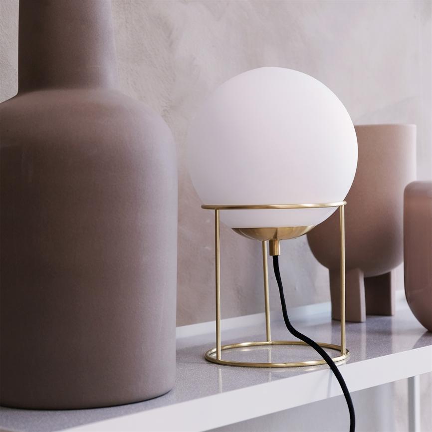 En hvid glaskugle-pære på en gylden metalsokkel står på en hvid hylde, kabel hænger ned; omgivet af matte, beige keramiske vaser foran en sandfarvet væg.