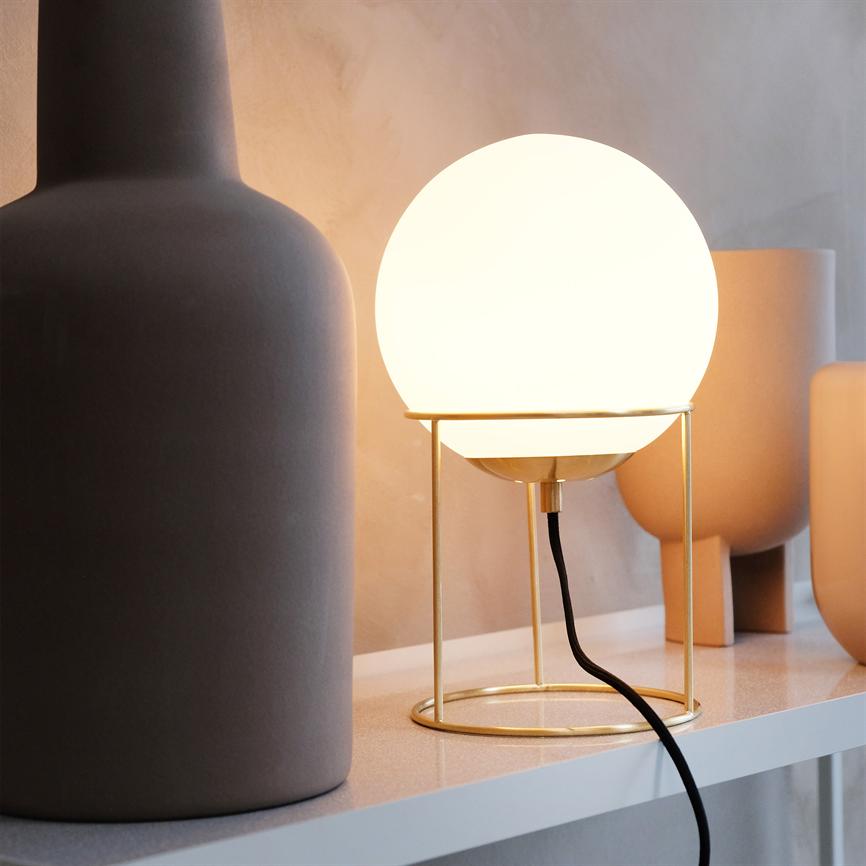 En rund lampe, der udsender varm, diffust lys, står i en gylden metalramme på en hylde omgivet af matte, neutrale keramikvaser.