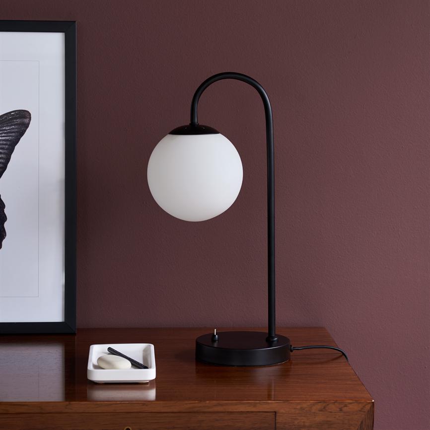 Sort metal bordlampe med hvid glasskærm hænger fra buet arm, står på træskuffe; ved siden af en hvid bakke med sæbe og kam; baggrund: bordeaux væg og indrammet plakat.