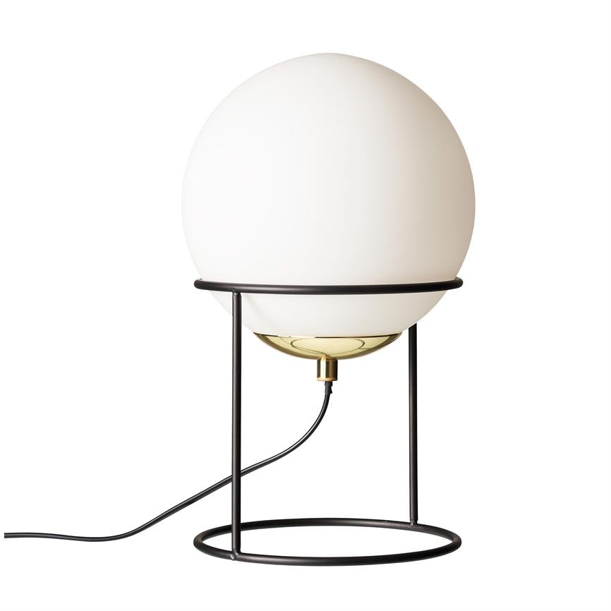 Rund opalglas-bordlampe hviler i en sort, cirkulær metalramme; glaskuppel omslutter en messing pæreholder, og en sort ledning fører ud mod venstre på hvid baggrund.