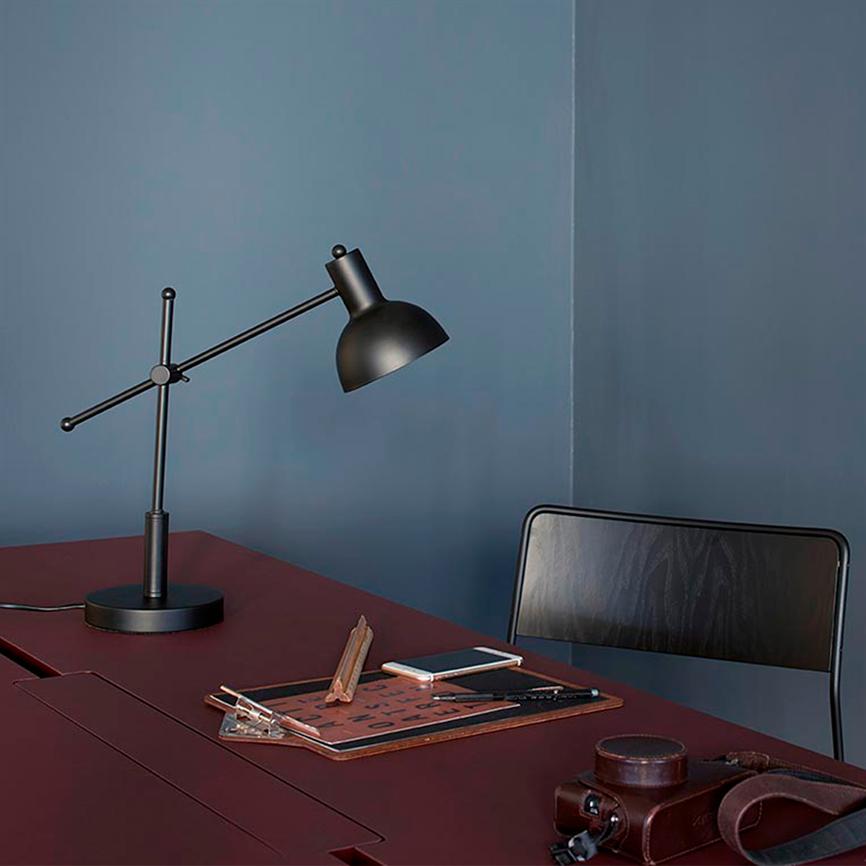 Sort skrivebordslampe peger nedad over et mørkerødt bord; en smartphone, notesblok, kuglepen og et brunt kamera ligger ved siden af, foran en mørk stol og en blå væg.