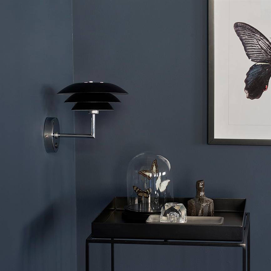 Sort væglampe monteret på en mørkblå væg; på et lille sort bord står en glaskuppel med tre monterede sommerfugle og en rustik keramisk figur ved siden af.