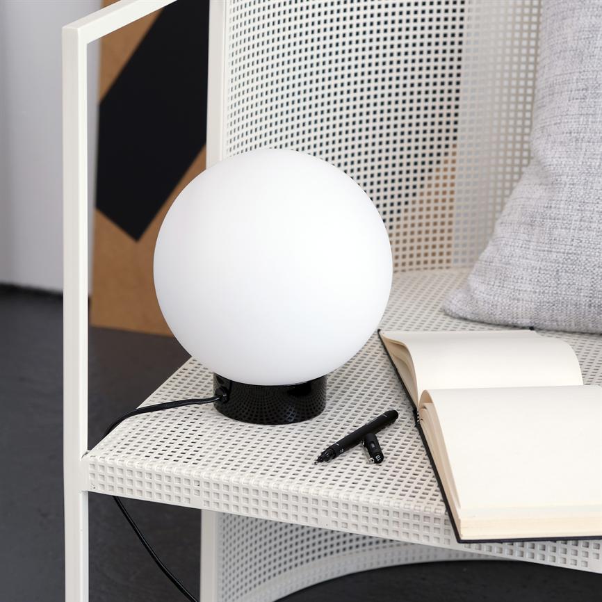 En rund hvid lampe står på en hvid perforeret hylde, lyser svagt; ved siden af ligger en åben notesbog og to sorte penne, med en grå pude i baggrunden.