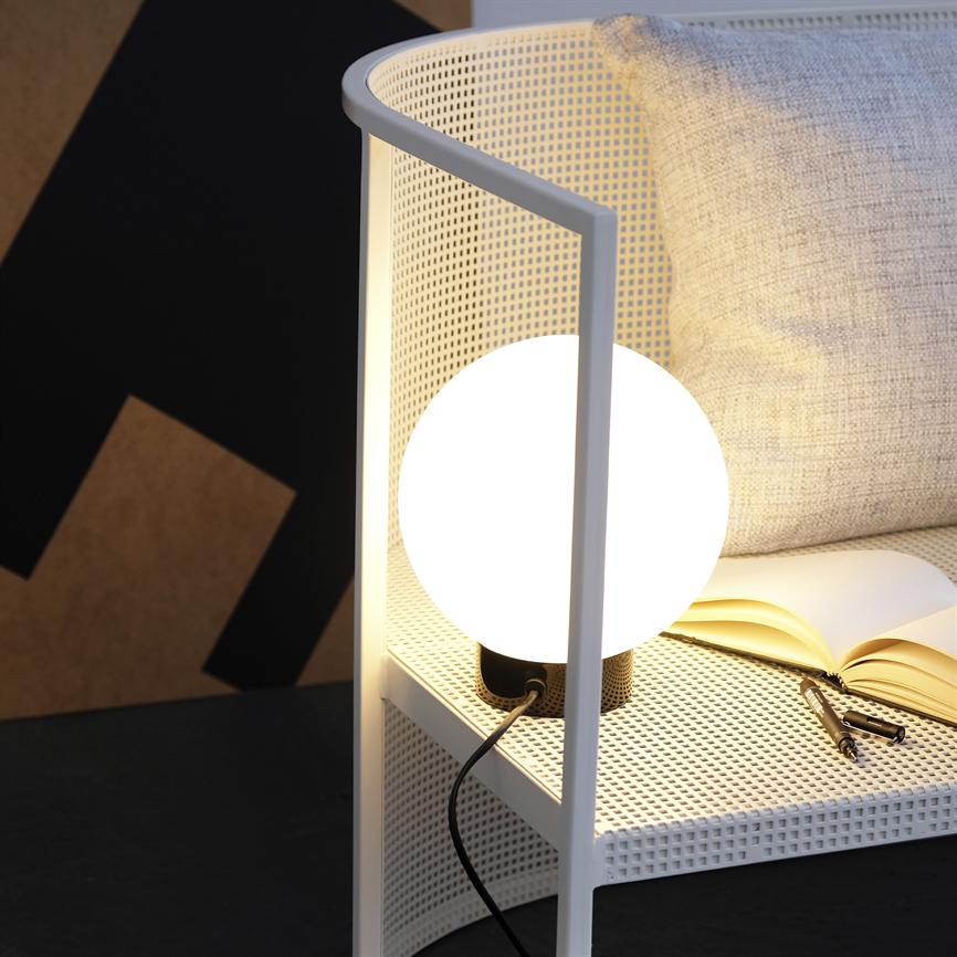 Rund bordlampe lyser varmt på et hvidt perforeret sidebord ved en lysegrå pude og en åben bog med pen, i et moderne, afdæmpet hjørne.