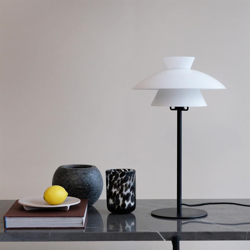 Hvid, lagdelt bordlampe står på sort sokkel; ved siden af ligger en indbundet bog med en hvid tallerken og en citron, samt to dekorative krukker på en grå marmorbordplade.