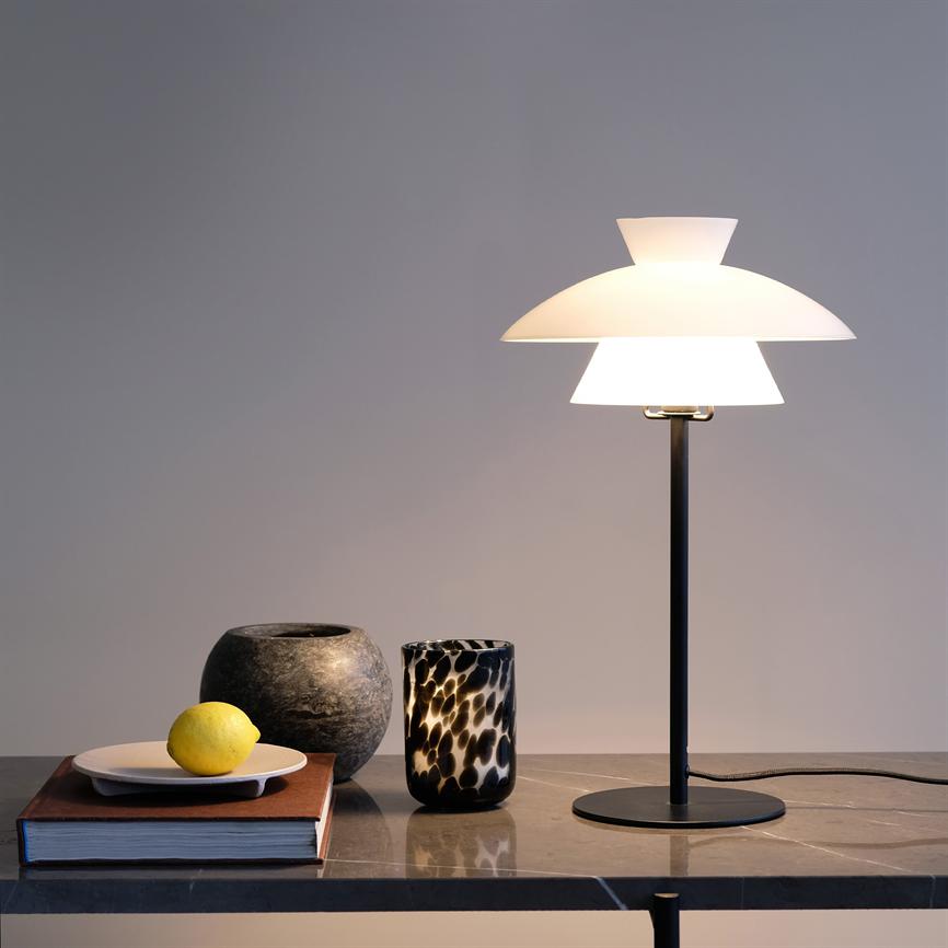 Bordlampe udsender blødt, varmt lys over et marmorbord; ved siden står en bog med en tallerken og citron, en rund stenvase og et sort-hvidt mønstret glas.