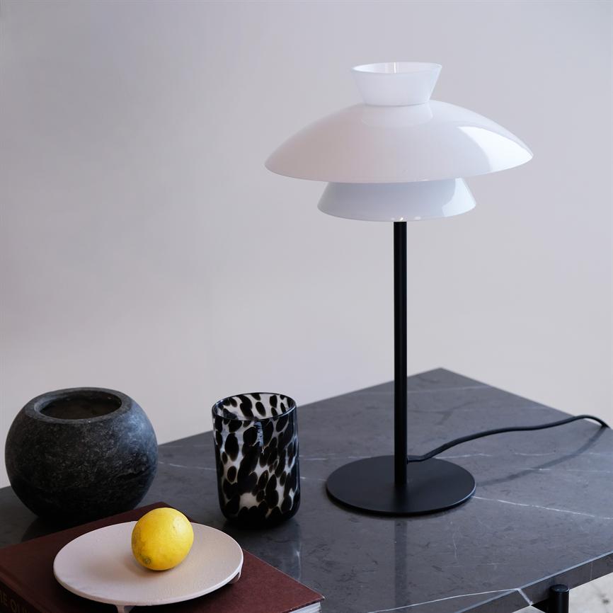 En hvid, moderne bordlampe står på et mørkt marmorbord; ved siden af ligger en gul citron på en lille tallerken, et mønstret glas og en rund stenskål.