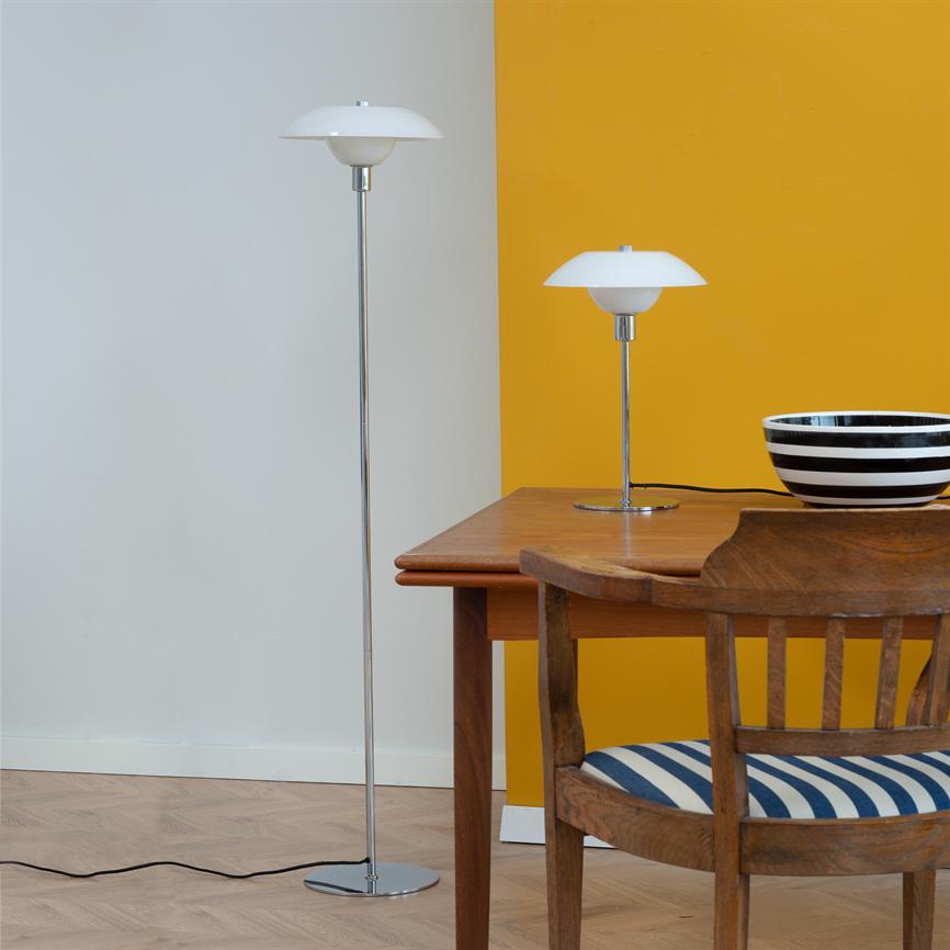 To hvide lamper står ved et træbord: en høj gulvlampe til venstre og en mindre bordlampe på bordet; en træstol med blå-hvid pude og en sort-hvid skål foran gul/lys væg.